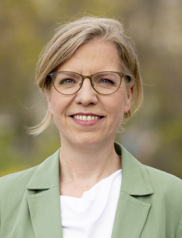 Leonore Gewessler speaking at a podium during the Green Party convention in Vienna, with delegates applauding in the background.