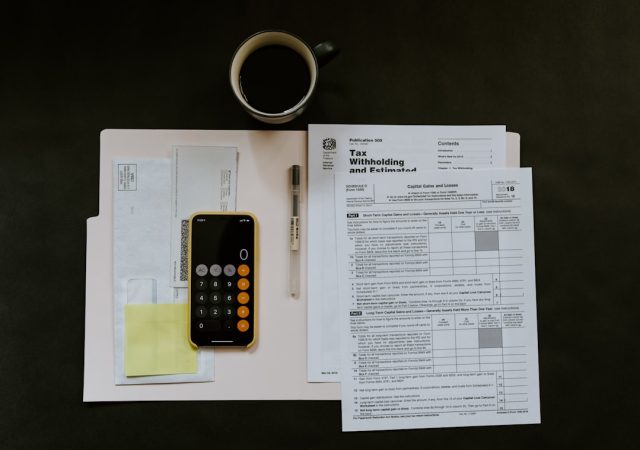 A stack of tax forms and a calculator on a desk, with a blurred office background suggesting a small business setting.